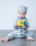 A baby sitting on a grey wood floor wearing a light blue and white camouflage print one-piece outfit and matching hat, holding a yellow toy.