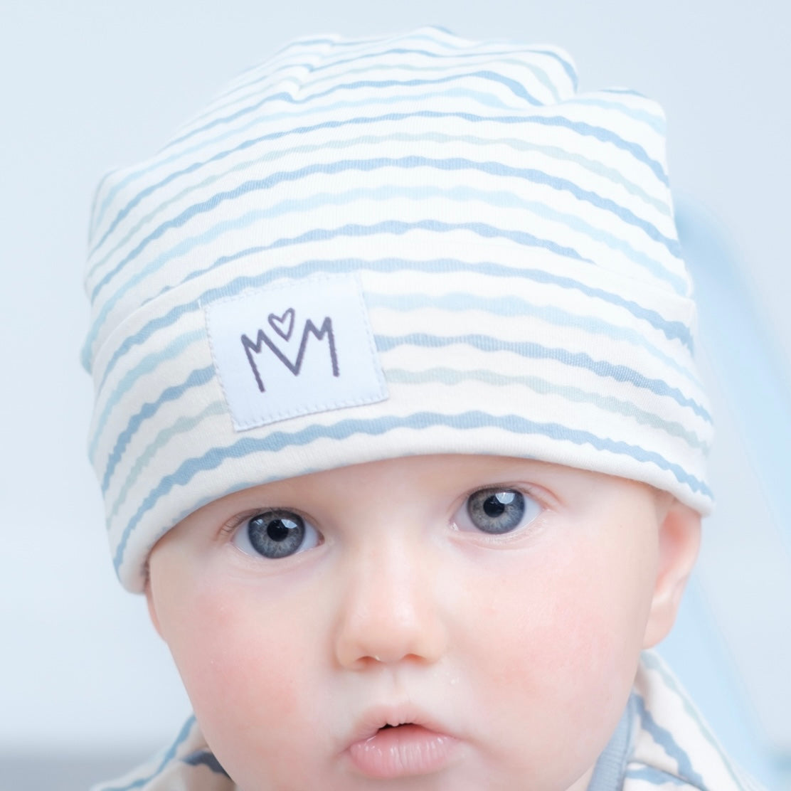 A close-up of a baby wearing a soft knit beanie hat with light blue and white horizontal stripes and a white patch featuring a black embroidered logo.