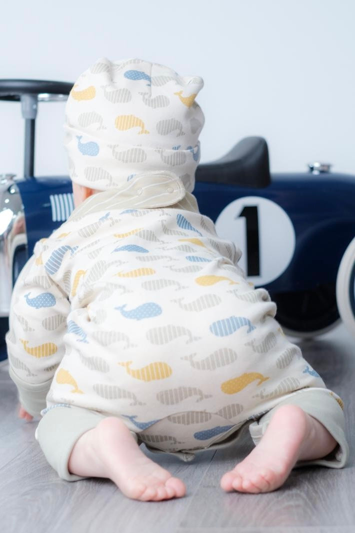 A baby crawling on a grey floor, wearing a cream-coloured hooded jumpsuit with a whale pattern in blue, yellow, and grey. The baby is positioned in front of a blue toy car.