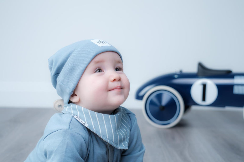 A baby wearing a light blue knit beanie and a matching blue and white striped bib, looking to the side with a toy race car blurred in the background.