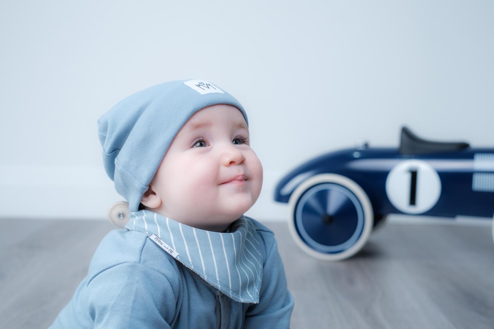 A baby wearing a light blue knit beanie and a matching blue and white striped bib, looking to the side with a toy race car blurred in the background.