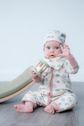 A baby sitting on a grey wooden floor wearing a cream-coloured patterned onesie and matching hat, holding a wooden toy rattle. A green and beige slide is visible in the background against a light grey wall.