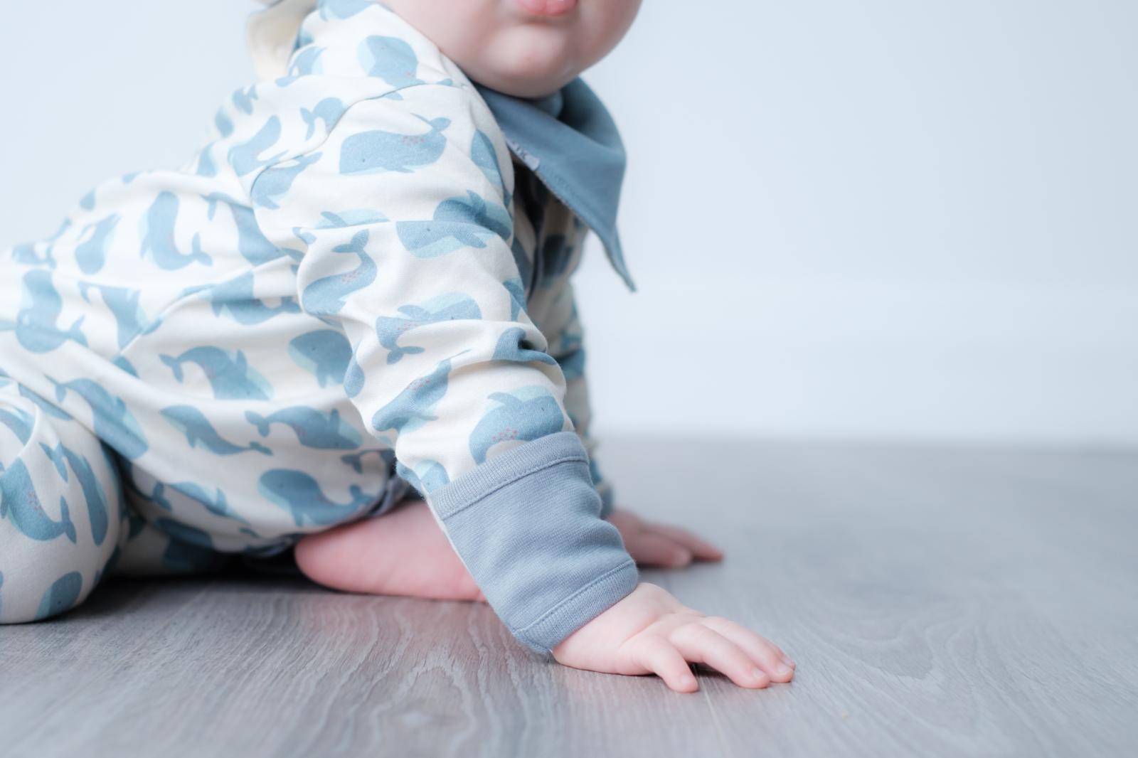 A baby crawling on a gray wood floor, wearing a light blue and white camouflage-patterned long-sleeve romper with a matching blue bib.