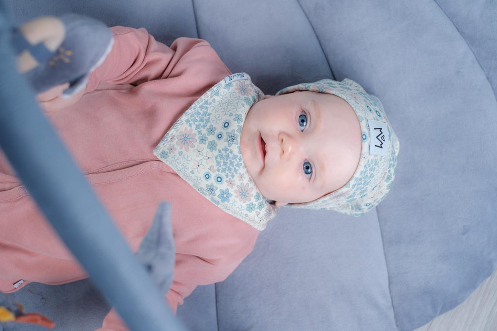 A baby lying on a blue padded surface, wearing a pink outfit, a light blue floral bib, and a matching light blue bonnet with the brand logo visible.