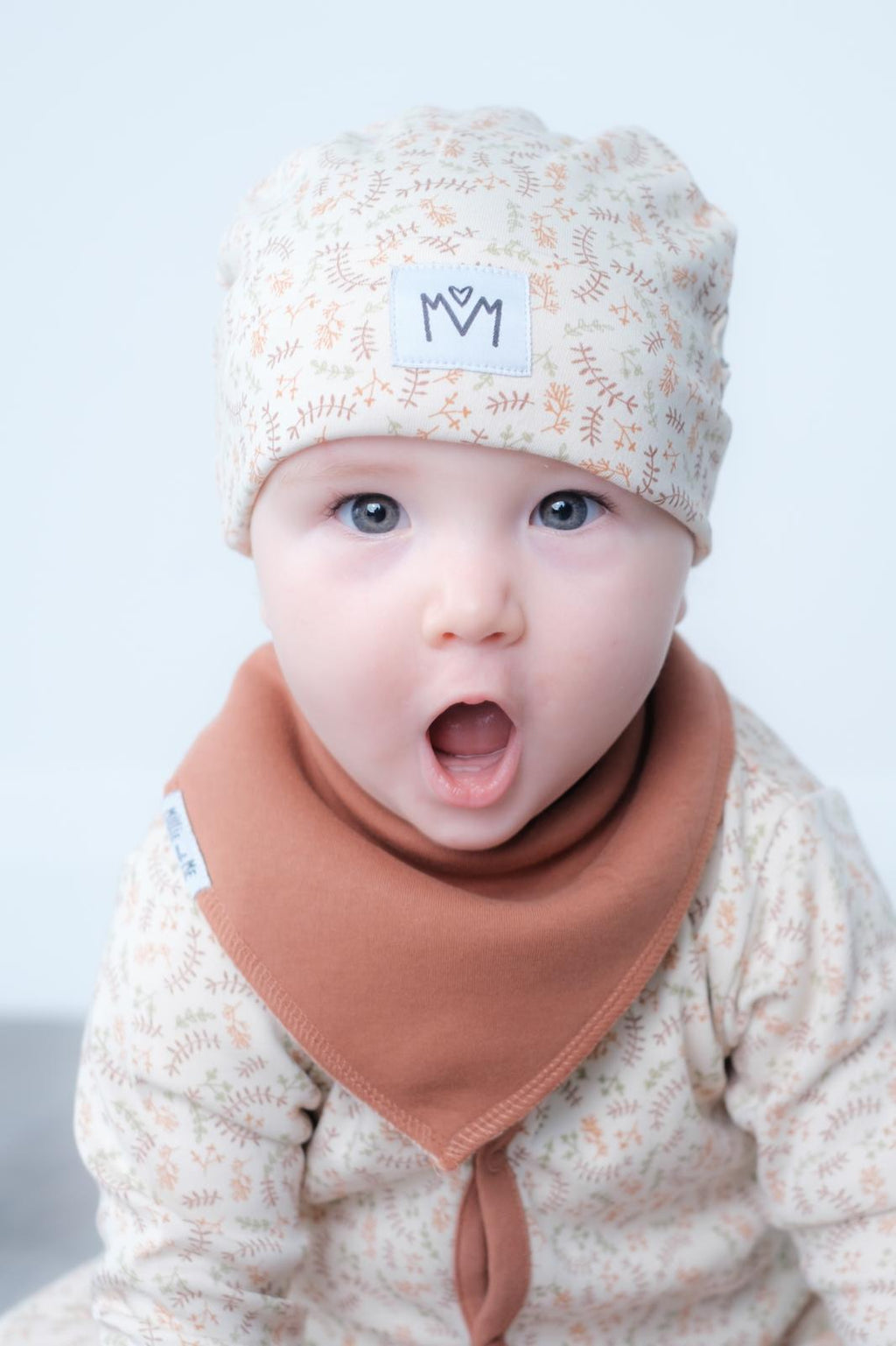 A close-up of a baby wearing a cream-colored patterned beanie with a small white logo patch and a terracotta-colored bandana bib, set against a plain light background.