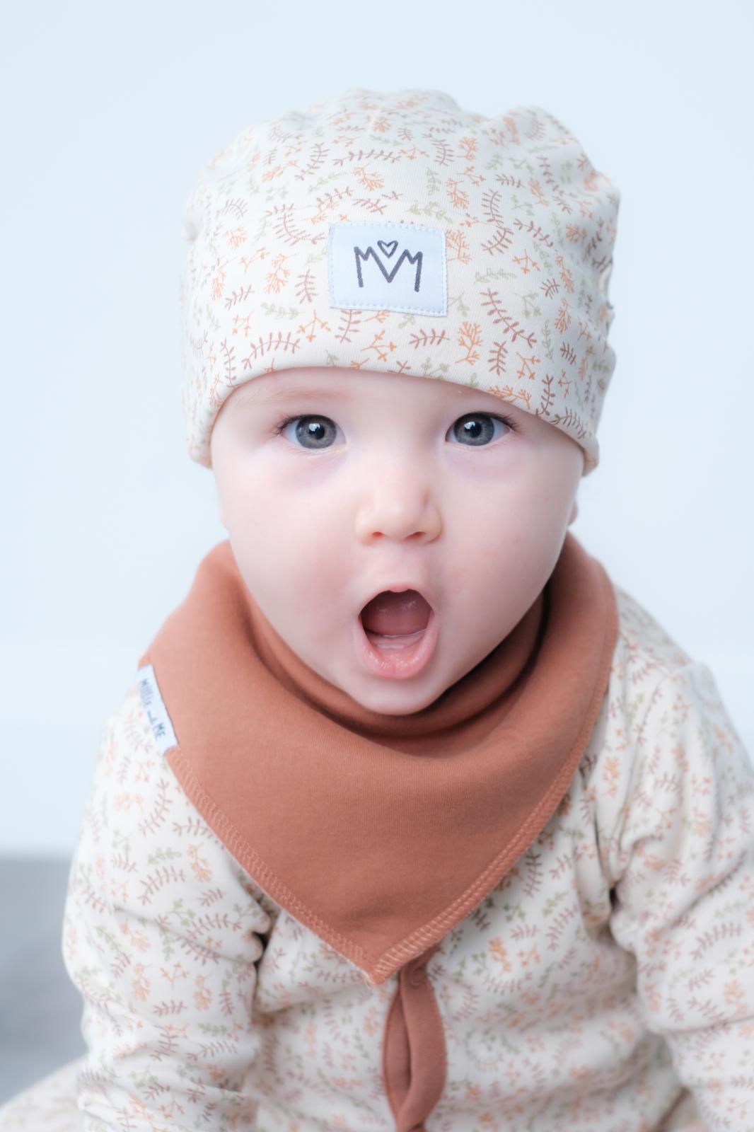 A close-up of a baby wearing a cream-colored patterned beanie with a small white logo patch and a terracotta-colored bandana bib, set against a plain light background.