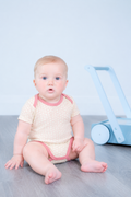 A baby sitting on a gray wooden floor wearing a cream short-sleeve bodysuit with pink trim and a subtle polka dot pattern.