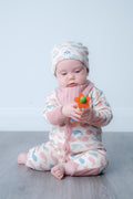 A baby sitting on a grey wooden floor against a light grey wall, wearing a pink and white patterned footless one-piece jumpsuit and matching beanie hat, holding a small orange and green toy.