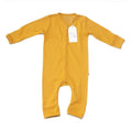 A mustard yellow long-sleeve baby footed sleeper with snap closures down the front, featuring a white hangtag with the brand name 'Millie and Me' printed on it, laid flat against a white background.