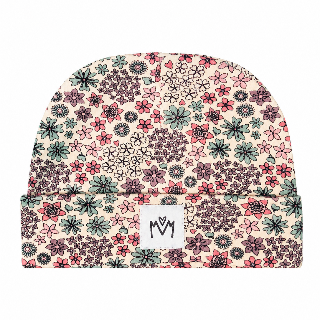 A folded knit beanie hat featuring an all-over dense floral and botanical pattern in pink, green, purple, and black on a light cream background. A small white rectangular brand patch with the stylised 'M M' logo is stitched onto the front fold.