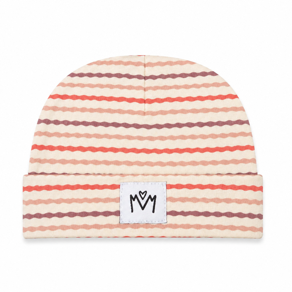 A cuffed knit beanie hat featuring horizontal wavy stripes in shades of coral red, dusty rose, and dark brown on a warm cream background. A small white rectangular patch on the front cuff displays the black hand-drawn logo 'MM'.