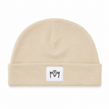 A beige knit beanie hat with a folded cuff, featuring a small white square patch on the front with a black embroidered 'M' logo.