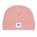 A dusty pink knit beanie hat with a folded cuff, featuring a small white square patch on the front with a black embroidered 'M' logo.