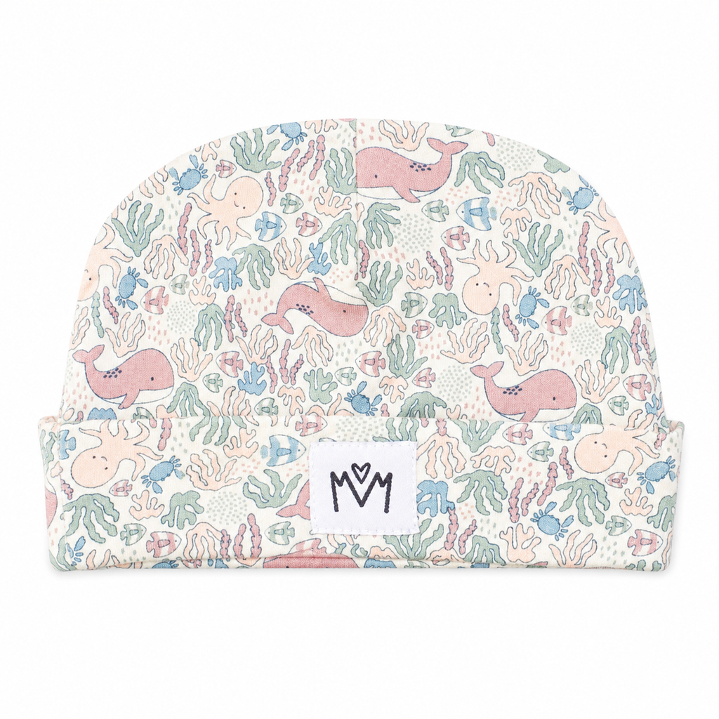A baby beanie hat featuring a pastel underwater print with pink whales, blue octopus, and green coral on a cream background, with a white brand patch reading 'M M' on the front cuff.