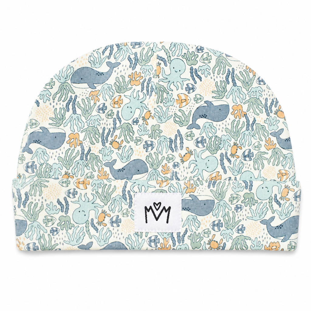 A baby beanie hat featuring an all-over underwater pattern with whales, octopus, fish, and sea plants in muted blue, teal, and orange on a cream background. A white fabric label with a black stylized 'M' logo is sewn onto the front.