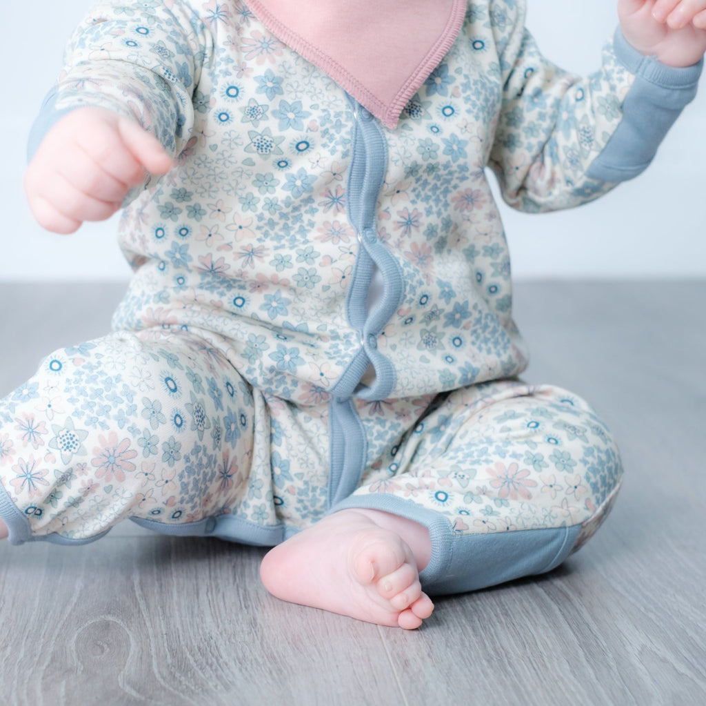 A baby sitting on a gray wood floor wearing a long-sleeve footless onesie with a light blue and pink floral pattern and a solid pink bib.