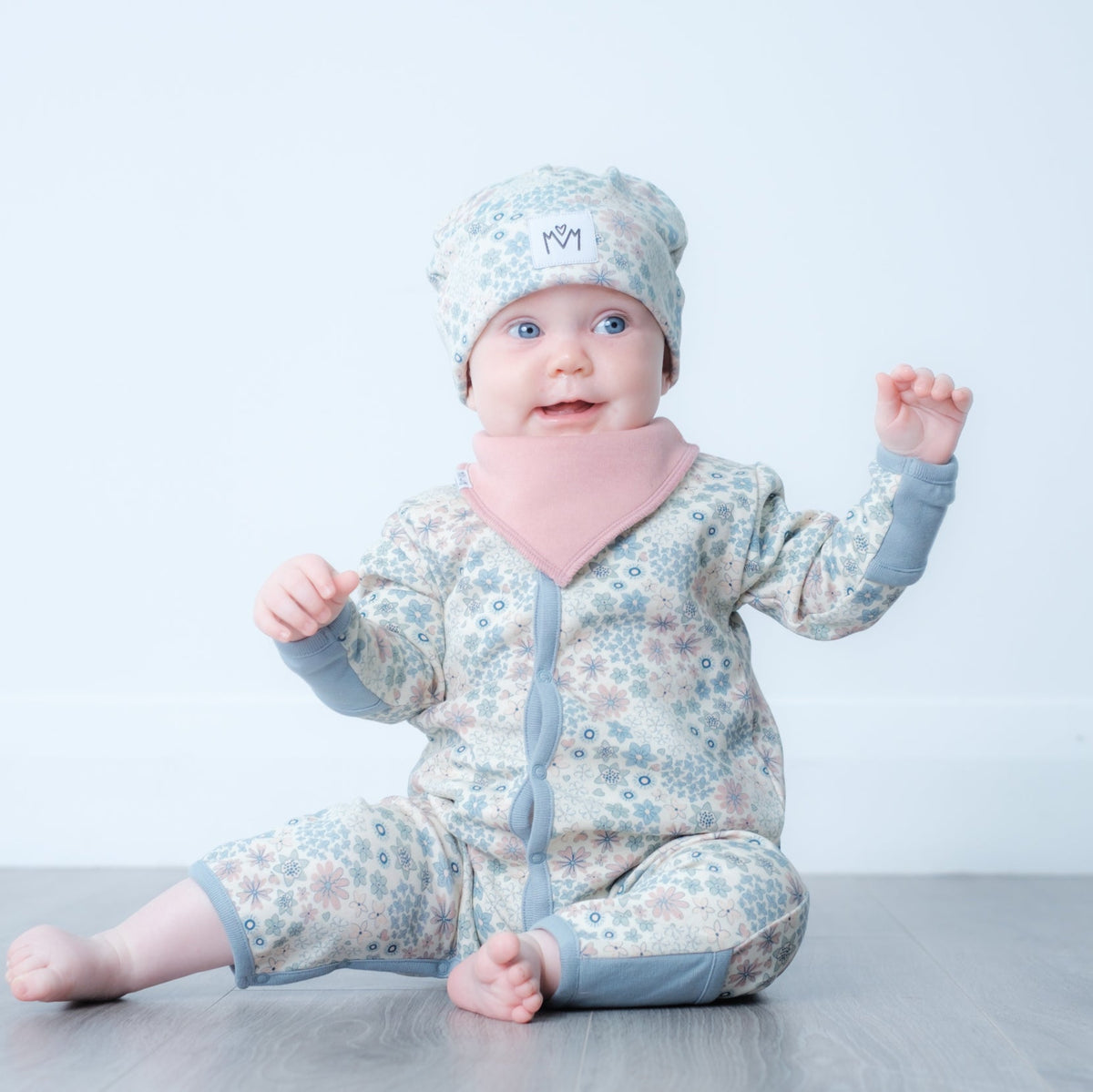 A baby sitting on a gray wooden floor against a white wall, wearing a light blue and white floral print one-piece outfit with a pink bib and matching hat.