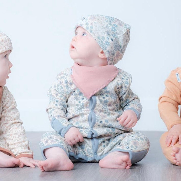 A baby sitting on a grey floor wearing a light blue and white floral print footie, a pink bib, and a matching floral bonnet. Two other babies are partially visible on either side.