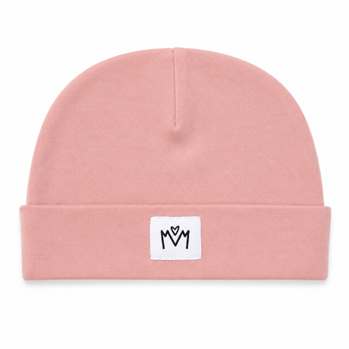 A dusty pink knit beanie hat with a folded cuff, featuring a small white square patch on the front with a black embroidered 'M' logo.