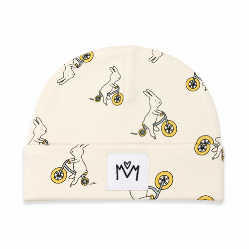 A cream-colored knit beanie hat featuring an all-over print of white rabbits riding yellow bicycles. A white rectangular patch on the folded cuff displays a black stylized 'MM' logo.