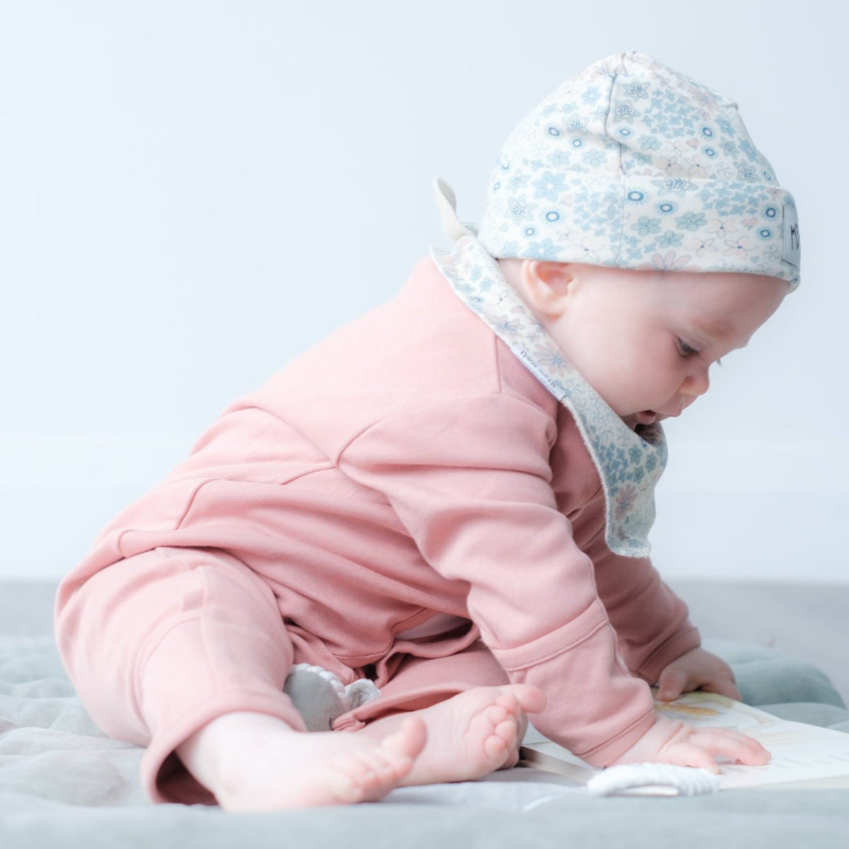 A baby wearing a pink outfit, a light blue floral bonnet, and a white bib sits on a light gray blanket on the floor, looking down at a book.