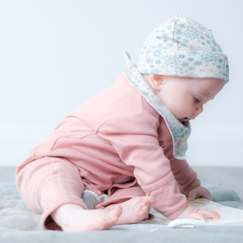 A baby wearing a pink outfit, a light blue floral bonnet, and a white bib sits on a light gray blanket on the floor, looking down at a book.