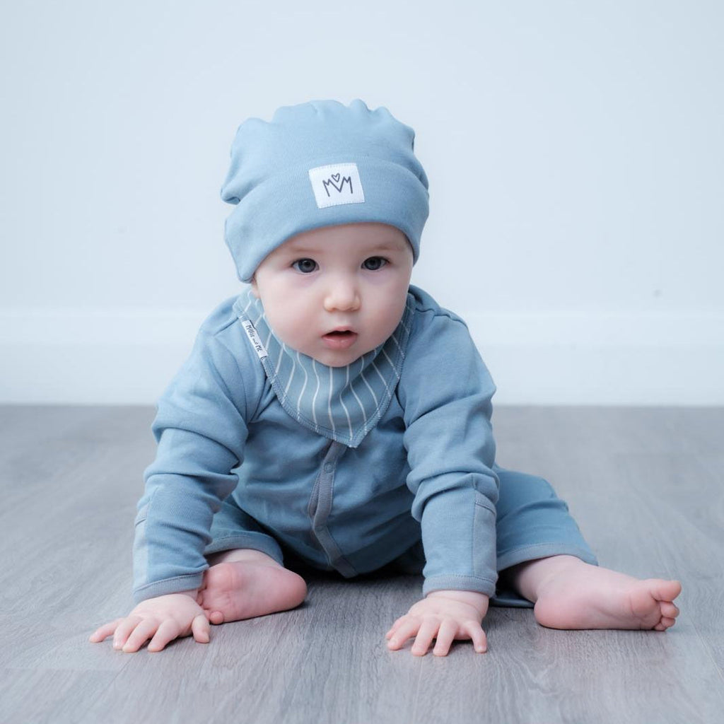 A baby crawling on a gray wooden floor against a light gray wall, wearing a light blue knit beanie with a small white square patch and a matching light blue long-sleeve outfit with a striped bib.
