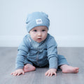 A baby crawling on a gray wooden floor against a light gray wall, wearing a light blue knit beanie with a small white square patch and a matching light blue long-sleeve outfit with a striped bib.