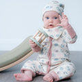 A baby sitting on a grey wooden floor wearing a cream-coloured patterned onesie and matching hat, holding a wooden toy rattle. A green and beige slide is visible in the background against a light grey wall.