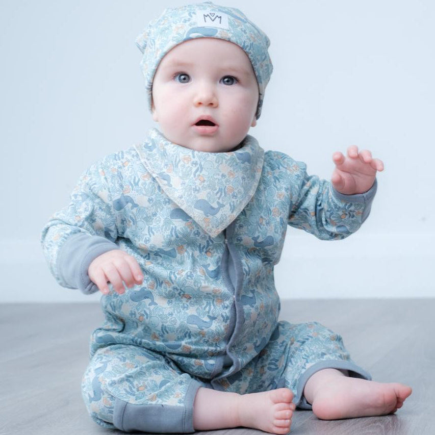 A baby sitting on a gray wood floor against a white wall, wearing a light blue patterned one-piece outfit with a matching hat and bib.