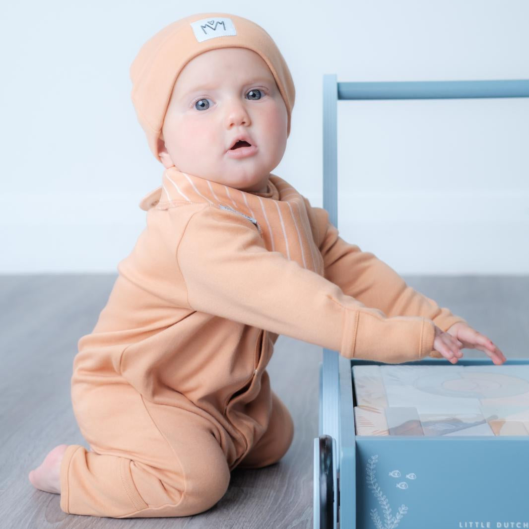 A baby wearing a peach-colored one-piece romper and matching beanie hat sits on a gray wood floor next to a light blue wooden toy cart.