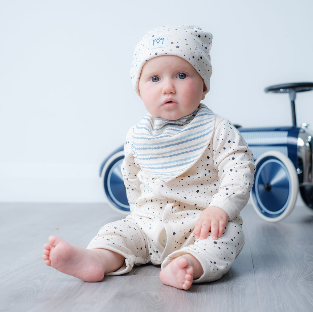 A baby sitting on a light gray floor wearing a cream-colored speckled one-piece outfit, a matching beanie, and a striped bib, with a blue and white toy car in the background.