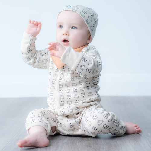 A baby sitting on a grey wood floor wearing a cream-coloured one-piece jumpsuit and matching bonnet with a small black geometric pattern, set against a plain light grey background.