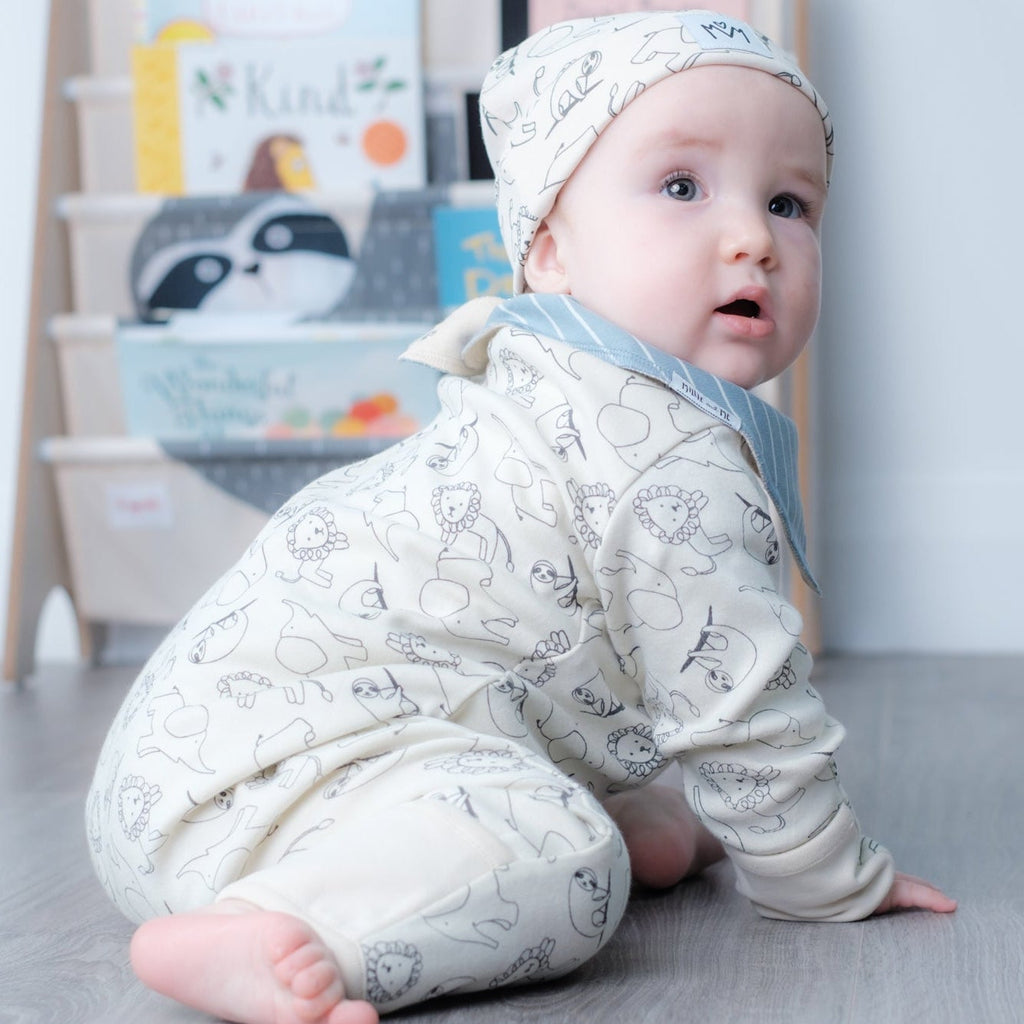 A baby wearing a cream-colored long-sleeve romper and matching beanie hat with a gray animal print, crawling on a gray wood-look floor in front of a light-colored bookshelf.