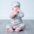 A baby sitting cross-legged on a gray wooden floor, wearing a light pink and blue ocean print one-piece outfit with a matching beanie hat.