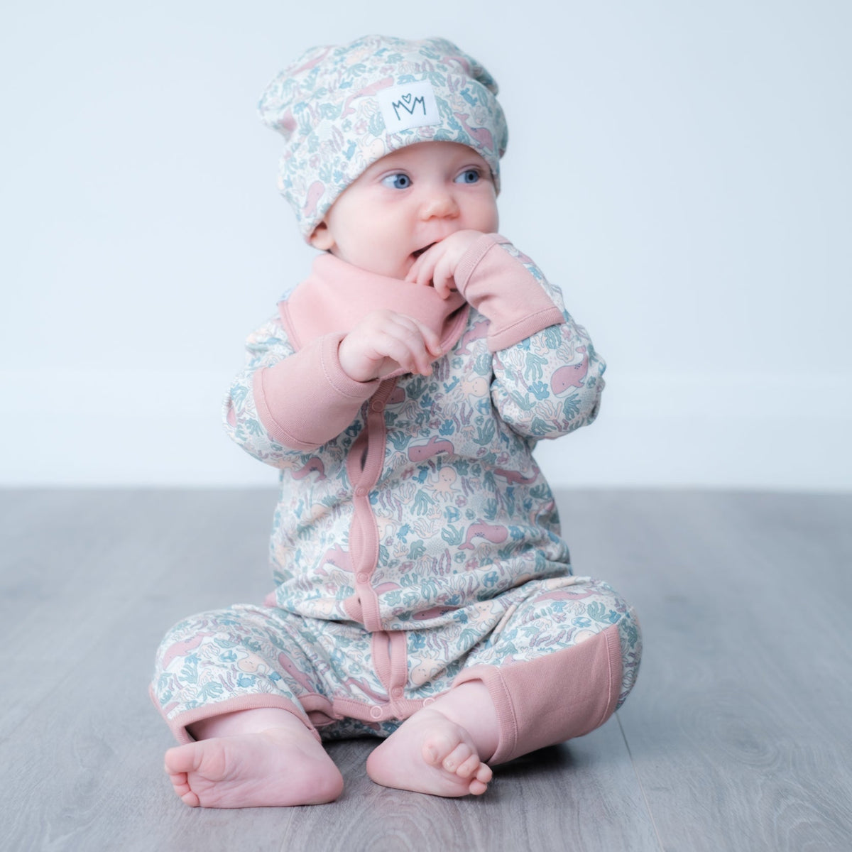 A baby sitting cross-legged on a gray wooden floor, wearing a light pink and blue ocean print one-piece outfit with a matching beanie hat.