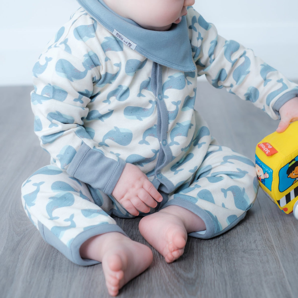 A baby sitting on a gray floor wearing a light blue and white whale-patterned button-up babygrow and matching hat, holding a yellow toy.