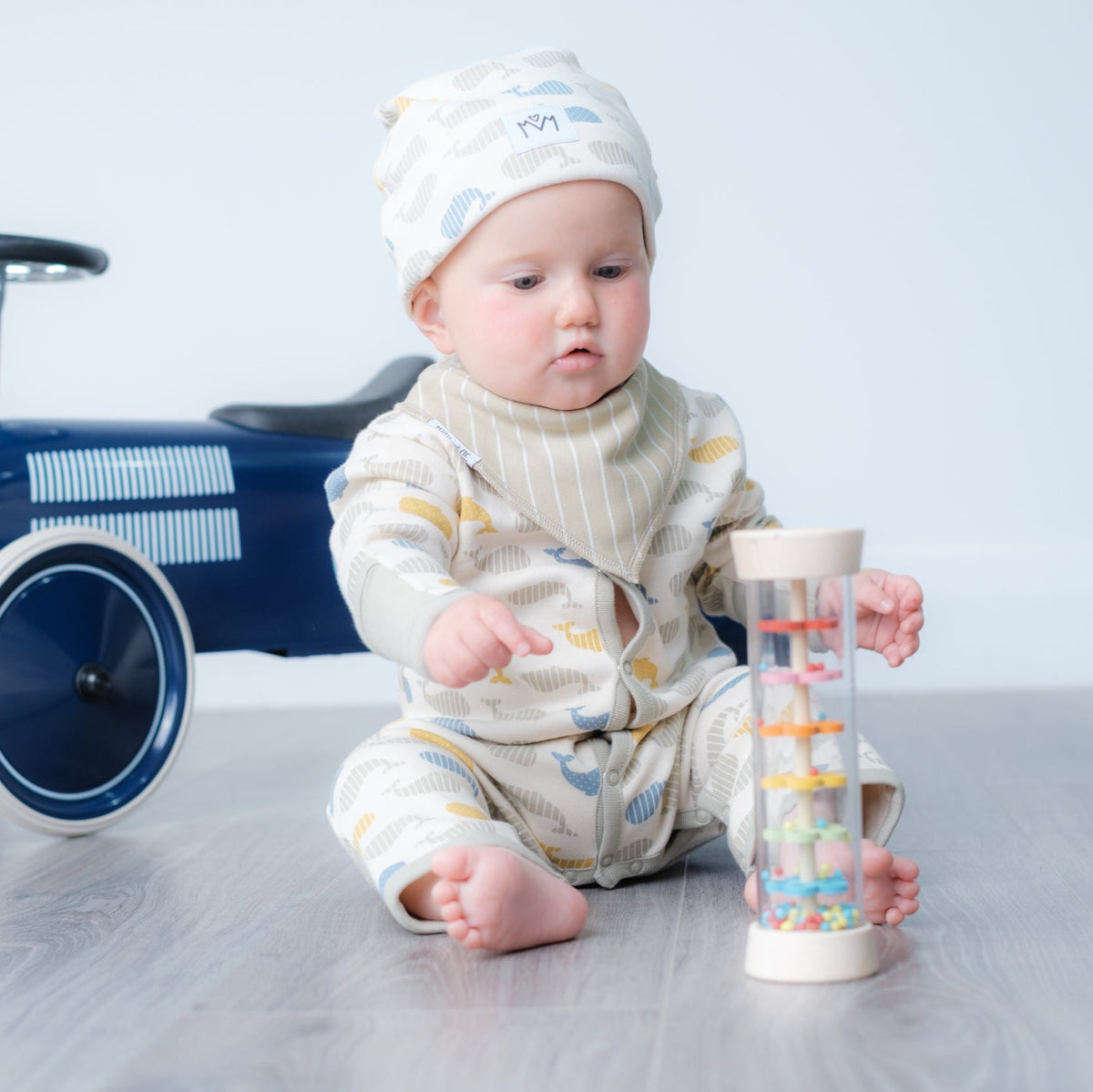 A baby wearing a cream-coloured striped onesie and matching hat sits on a gray floor next to a blue ride-on toy car, holding a clear plastic toy with colourful rings.