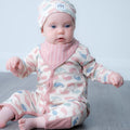 A baby wearing a matching long-sleeve footless romper and hat set with a pink and blue whale pattern on a cream background, sitting on a grey floor against a light blue wall.