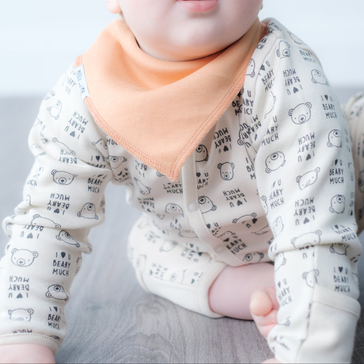 A baby wearing a cream-colored long-sleeve onesie with a black bear print, a matching beanie, and a peach-colored bib, lying on a light gray surface.