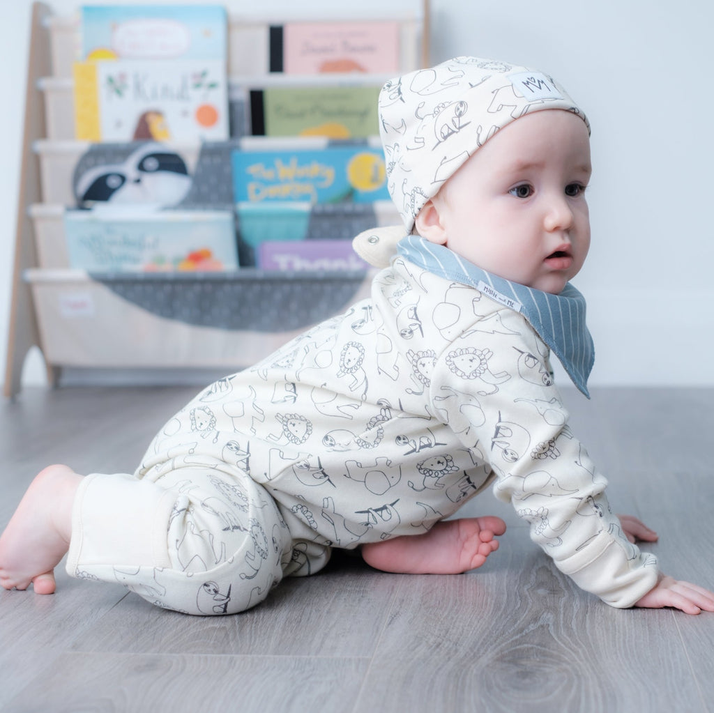 A baby crawling on a gray wood floor, wearing a cream-colored long-sleeve onesie and matching bonnet with a light blue striped bib.