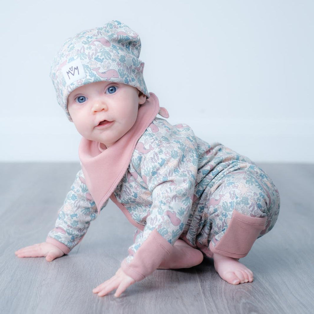 A baby crawling on a grey floor against a light grey background, wearing a floral print one-piece outfit with dusty pink accents and a matching beanie hat.