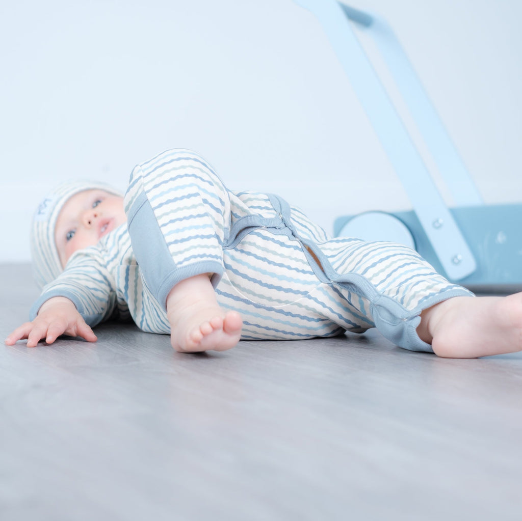 A baby lying on their back on a gray wooden floor, wearing a light blue and white striped footless one-piece outfit with a matching hat. A light blue toy walker is visible in the background against a white wall.