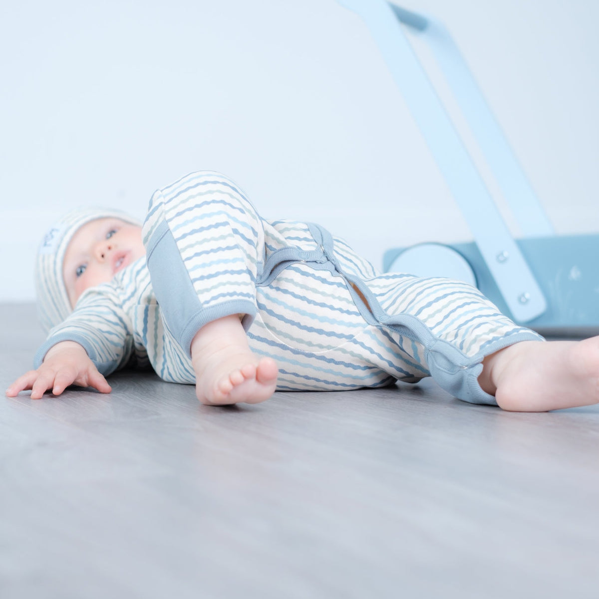 A baby lying on their back on a gray wooden floor, wearing a light blue and white striped footless one-piece outfit with a matching hat. A light blue toy walker is visible in the background against a white wall.