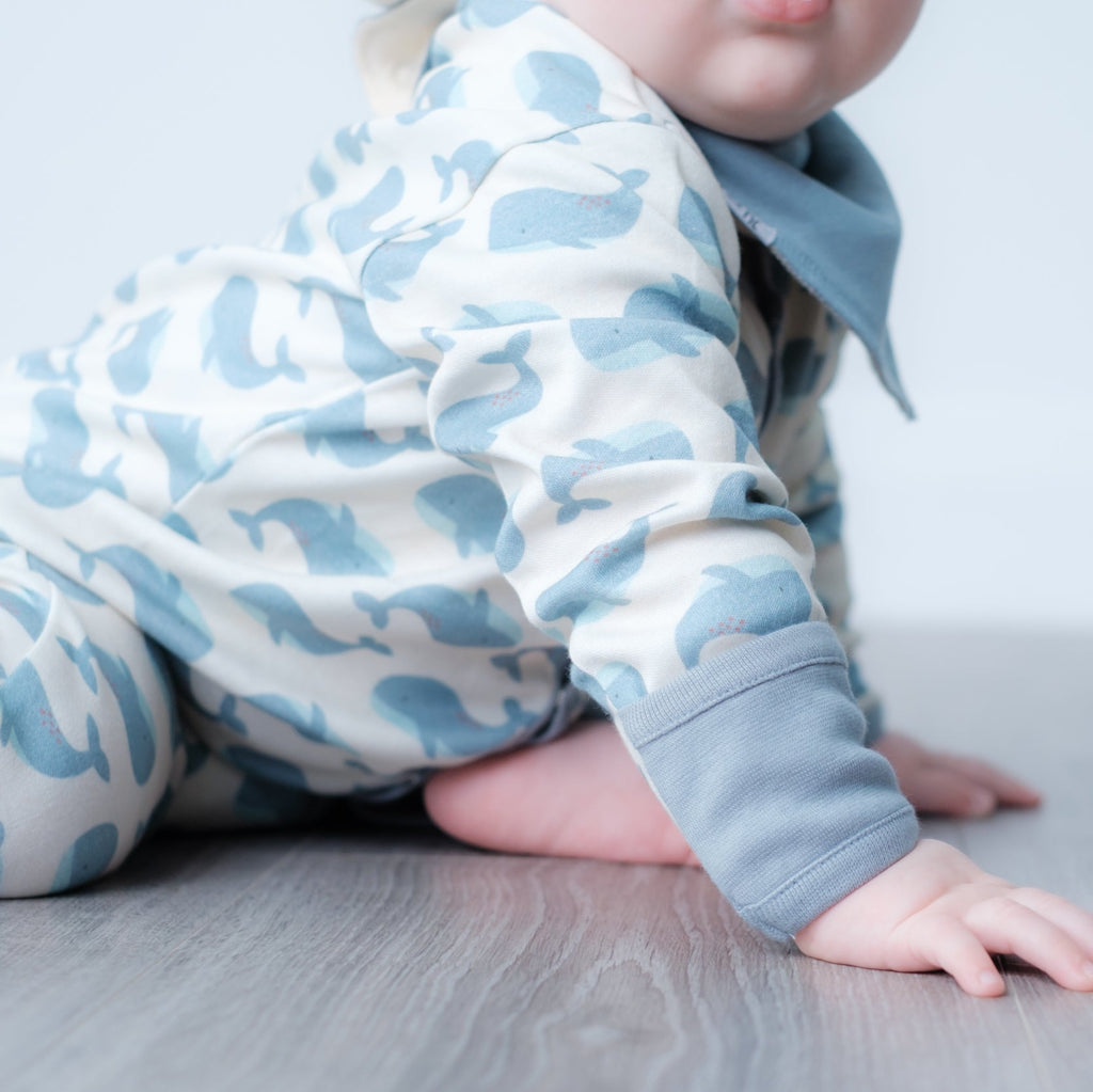 A baby crawling on a gray wood floor, wearing a light blue and off-white whale-patterned long-sleeve romper with a matching blue bib.