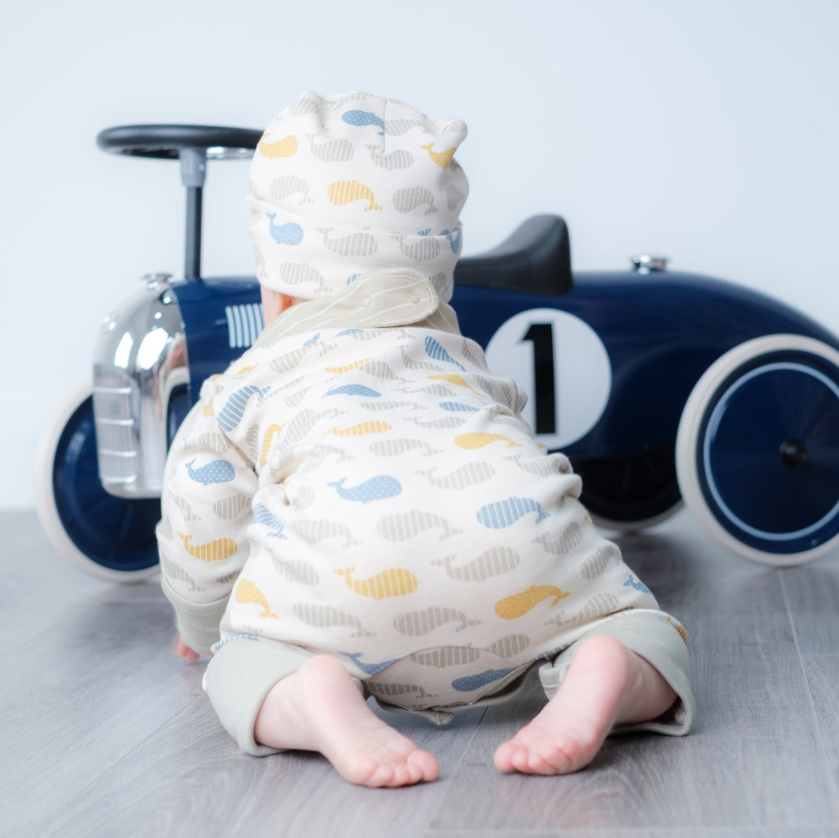 Baby wearing a cream organic cotton hooded romper with a blue, yellow and grey whale print, sitting in front of a vintage blue toy car.