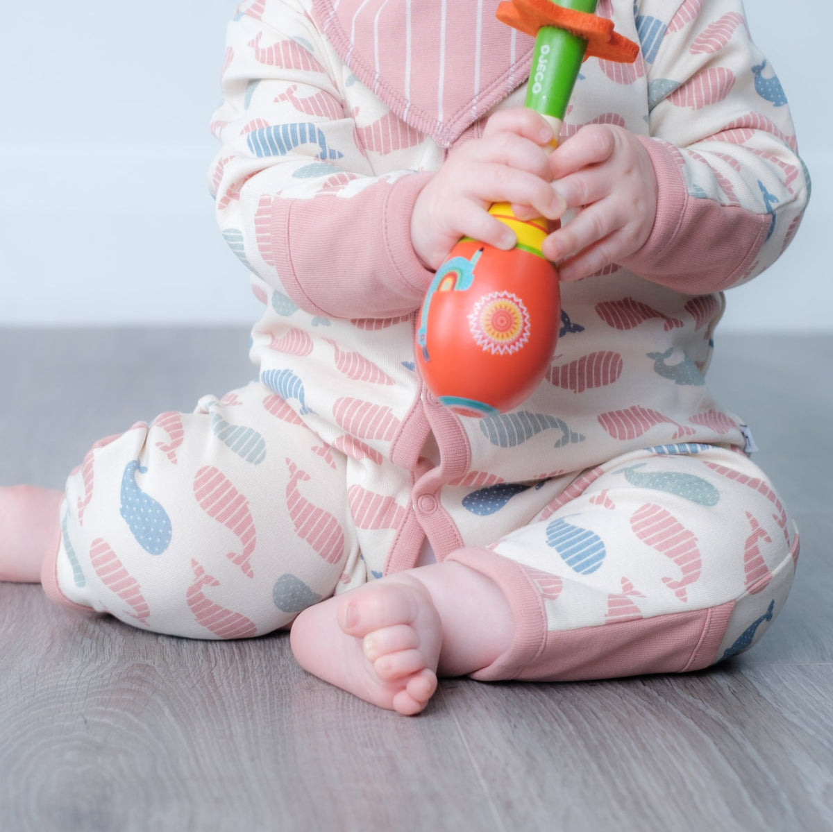 A baby sitting on a gray wood floor wearing a pink and blue patterned romper, holding a colourful toy.