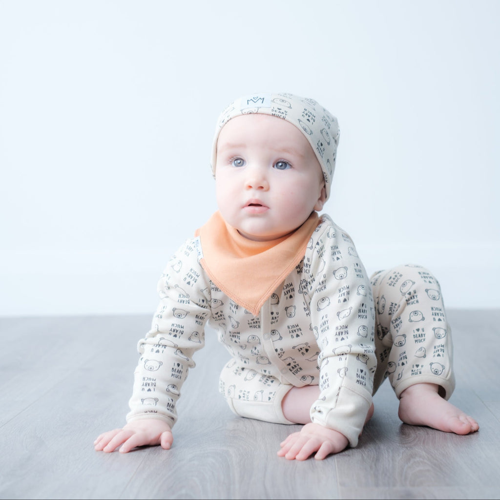 A baby crawling on a gray floor against a white studio background, wearing a cream-colored patterned long-sleeve romper, matching beanie, and an orange bib.