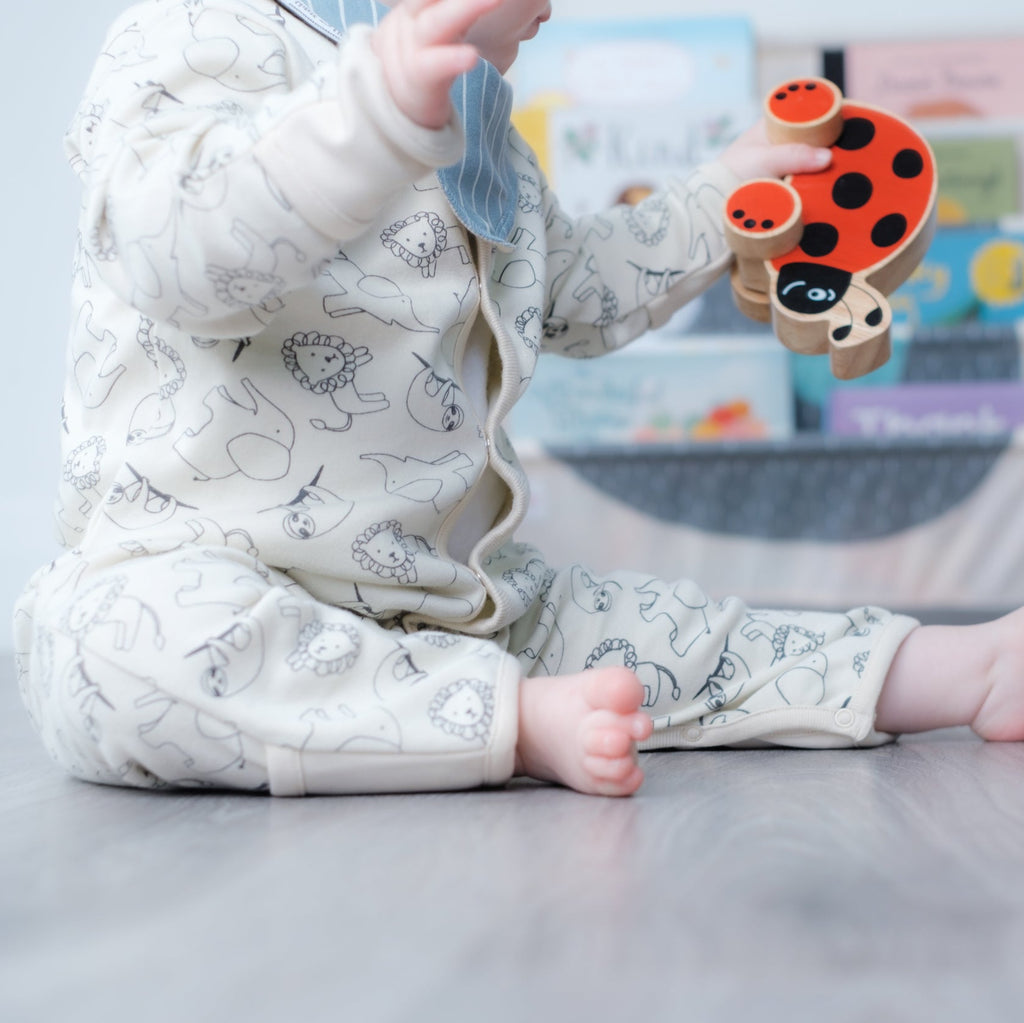 A baby sitting on a gray floor wearing a cream-colored onesie with a jungle pattern, holding a wooden ladybug toy in a nursery setting.