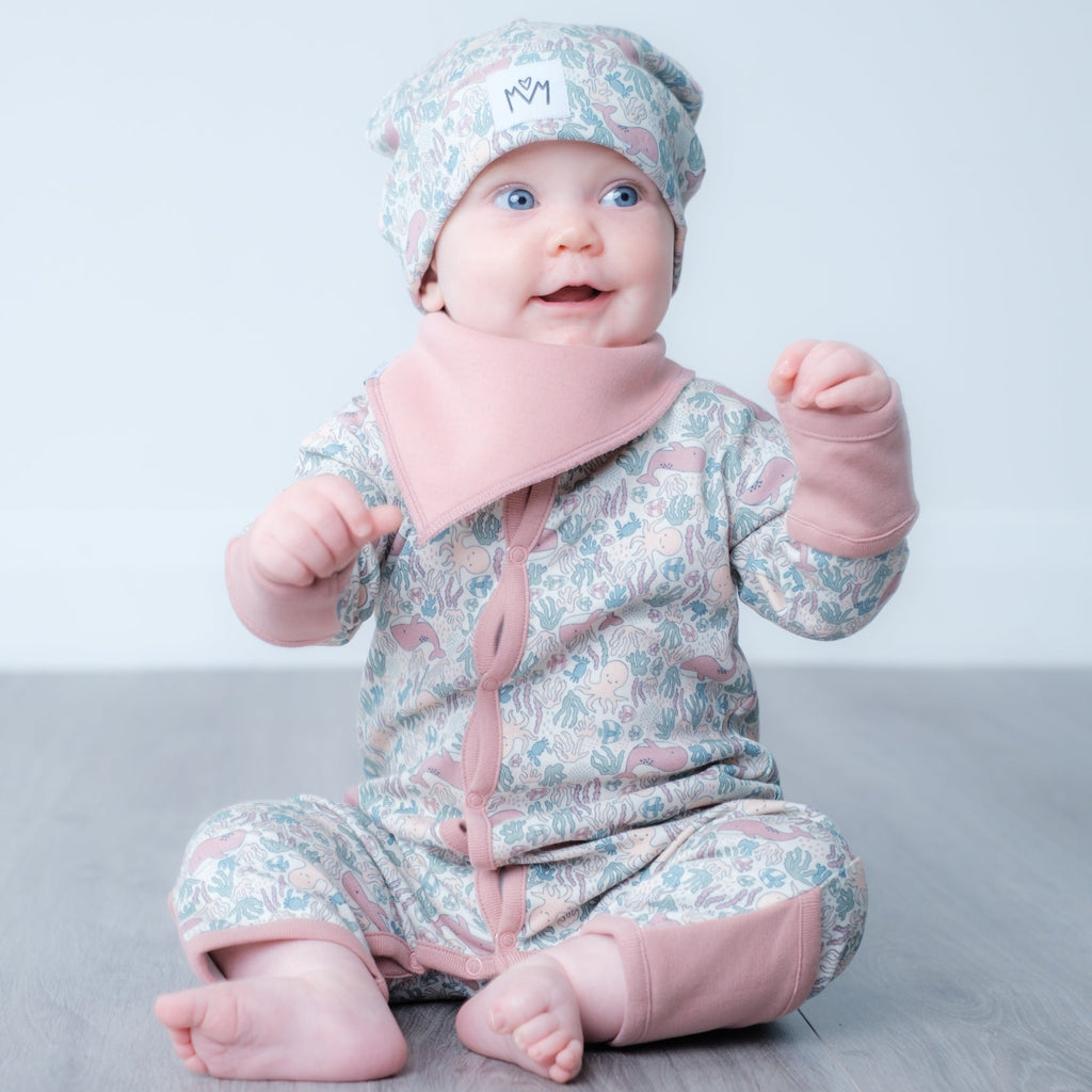 A baby sitting on a grey wooden floor wearing a light blue and pink ocean patterned footed babygrow with a matching bonnet and a pink bib.